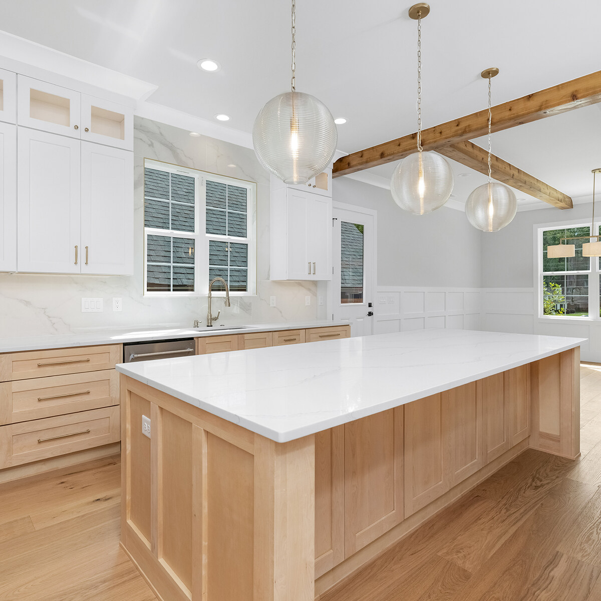 White kitchen with a full-length quartz-topped light wood island, three glass globe pendants, exposed wood beam, and marble backsplash